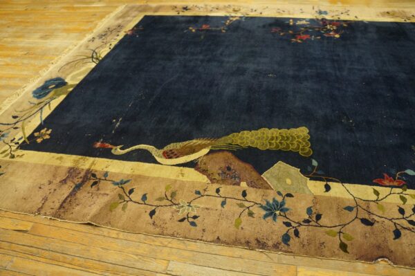 A high-pile navy blue rug featuring a large peacock motif and detailed tan floral borders rests on wood floors.