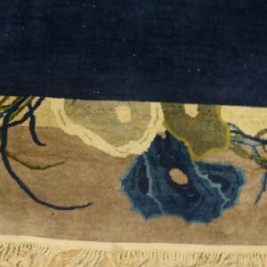 Thick pile rug detail showing a navy top border, tan field, blue floral design, and cream fringe.