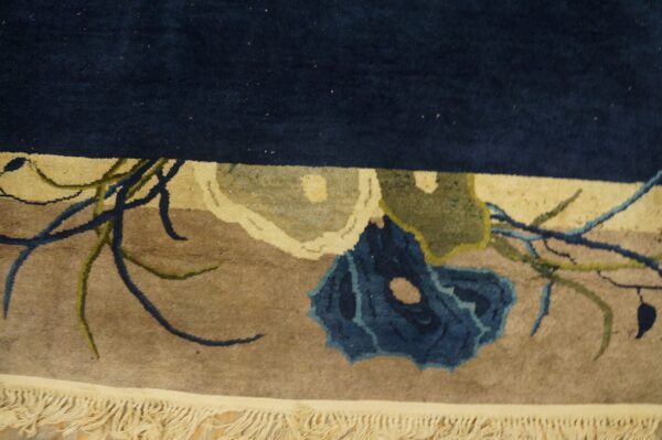 Thick pile rug detail showing a navy top border, tan field, blue floral design, and cream fringe.