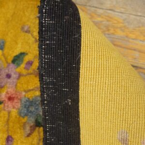 High-pile yellow floral rug edge curled to show dark binding and ribbed yellow backing on wood.