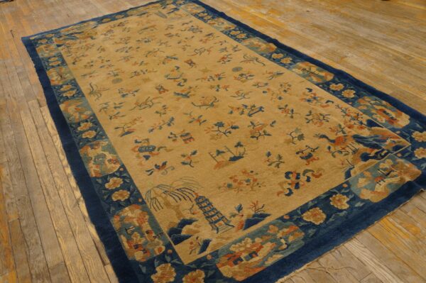 A tan rug with scattered blue and rust pictorial designs and a wide navy border rests on distressed wooden floorboards.