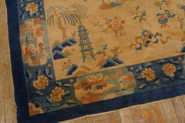 Tan rug with blue border and detailed scenic pattern featuring pagodas and trees on wood floors.