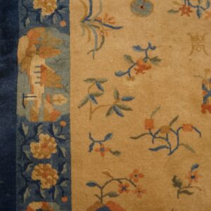 Tan low-pile rug with blue and orange botanical motifs and a dark blue border on wood flooring.