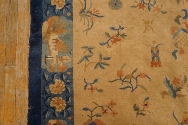 Tan low-pile rug with blue and orange botanical motifs and a dark blue border on wood flooring.