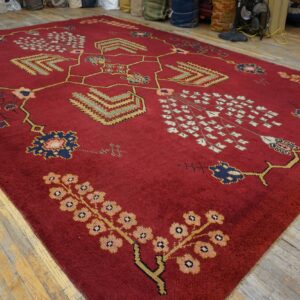 Deep red, medium pile rug with intricate gold, blue, and pink patterns on distressed wood flooring.