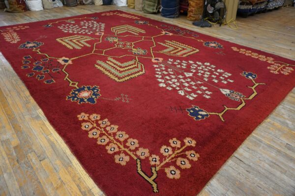 Deep red, medium pile rug with intricate gold, blue, and pink patterns on distressed wood flooring.