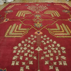 Stylized red rug with geometric yellow, blue, and cream motifs displayed on light wood flooring.