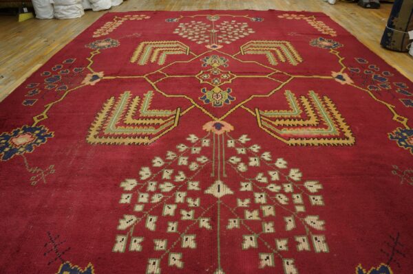 Stylized red rug with geometric yellow, blue, and cream motifs displayed on light wood flooring.