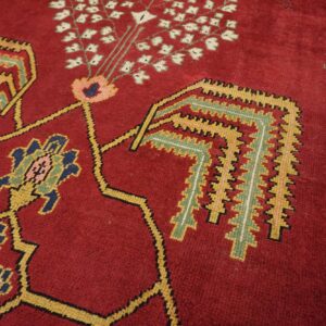 Angled close-up of a deep red rug with traditional geometric motifs in yellow, green, pink, and navy blue.