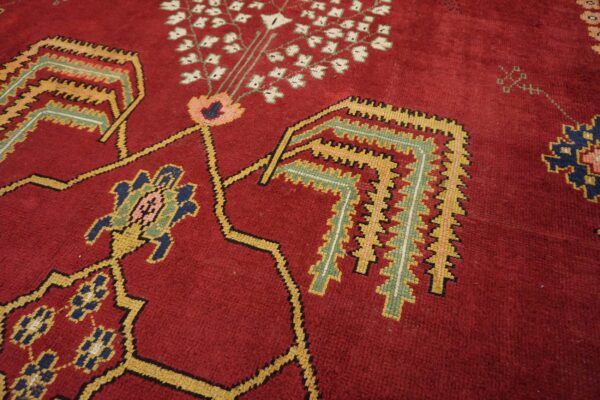 Angled close-up of a deep red rug with traditional geometric motifs in yellow, green, pink, and navy blue.