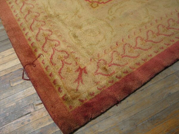A high-pile beige rug with a red abstract border and motif rests on distressed wooden floorboards.