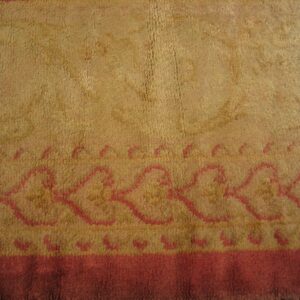 Textured camel-colored rug with a pink outline border featuring a scalloped geometric pattern and solid red band.