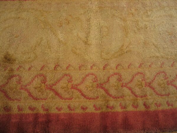 Textured camel-colored rug with a pink outline border featuring a scalloped geometric pattern and solid red band.