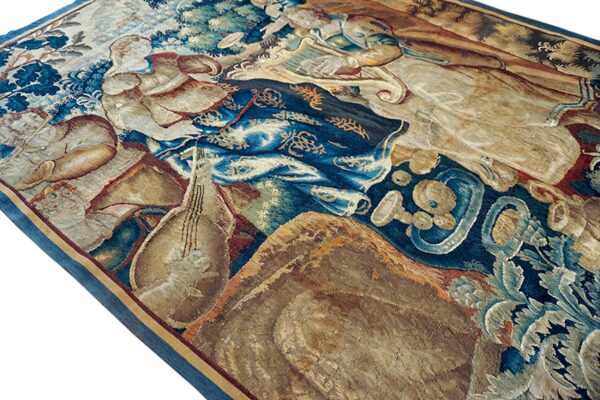 Detail of a tapestry-style rug showing figures and foliage woven in deep blue, beige, and warm brown colors.