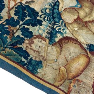 Close-up showing aged, flat-woven textile with figures, dark teal foliage, and a distinct blue fabric edge border.
