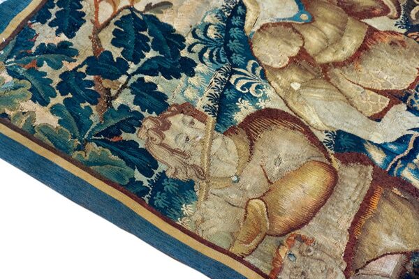 Close-up showing aged, flat-woven textile with figures, dark teal foliage, and a distinct blue fabric edge border.