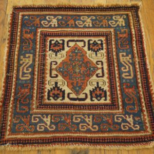 Small, patterned rug in blue, rust, and tan tones with short fringe on rustic wood planks.