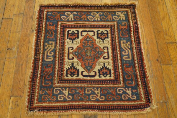 Small, patterned rug in blue, rust, and tan tones with short fringe on rustic wood planks.