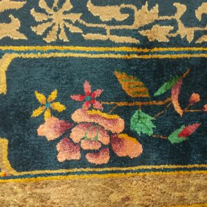 Detail of a plush rug showing a teal field with coral flowers framed by tan and gold scrollwork.