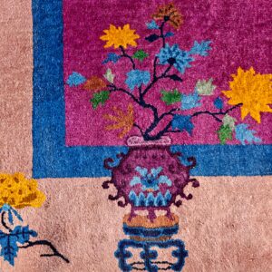 Pinkish-beige rug corner with magenta and blue borders surrounding a floral pattern in a decorative vase.