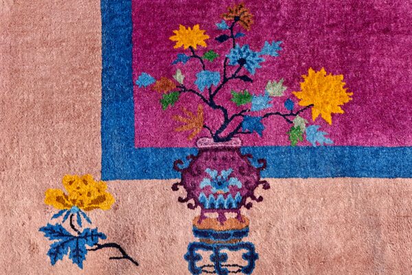 Pinkish-beige rug corner with magenta and blue borders surrounding a floral pattern in a decorative vase.