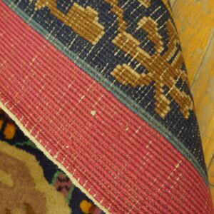 Rolled rug edge showing pink flat binding, a geometric blue and brown border, and yellow distressed wood flooring.