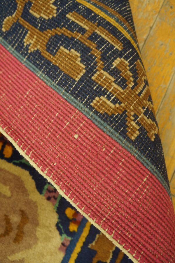 Rolled rug edge showing pink flat binding, a geometric blue and brown border, and yellow distressed wood flooring.