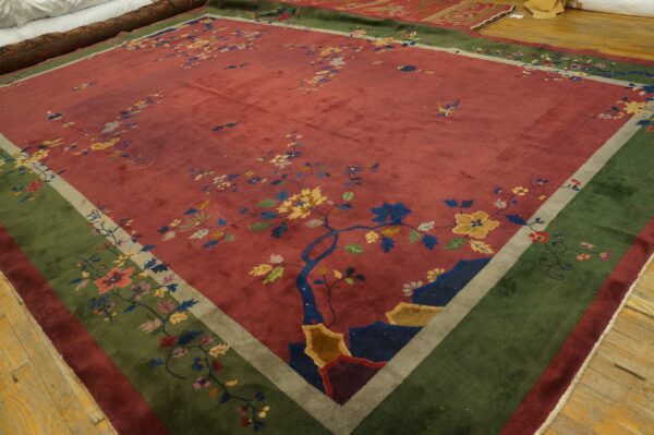 Multicolored floral rug with a deep red field and broad olive green border rests on light wood flooring.