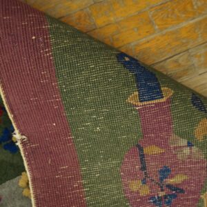Woven rug showing burgundy border and green floral pattern, curled on distressed yellow plank flooring.