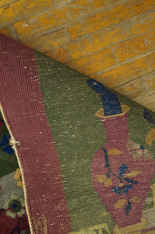 Woven rug showing burgundy border and green floral pattern, curled on distressed yellow plank flooring.
