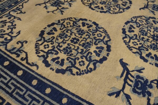 A detailed low-pile rug features dark and light blue medallions and a key pattern on a beige field.