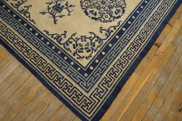 Cream rug with detailed indigo blue floral patterns and geometric meander borders on old wood plank flooring.