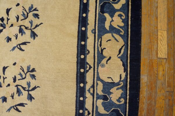 A tan rug with blue floral sprays and a detailed wide border lies on worn wooden flooring.