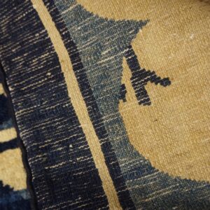 Coarse-textured rug closeup with dark blue, faded blue, and tan geometric patterns over wood.