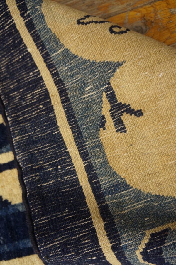 Coarse-textured rug closeup with dark blue, faded blue, and tan geometric patterns over wood.