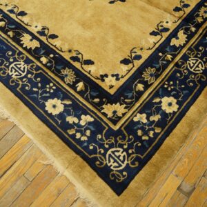 Gold-tan rug corner with a thick navy border patterned with cream flowers and symbols on worn wood floorboards.