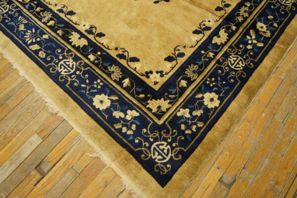 Gold-tan rug corner with a thick navy border patterned with cream flowers and symbols on worn wood floorboards.