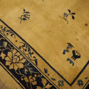 Gold rug corner showing a dark blue floral border and scattered stylized motifs in the field over wood floor.