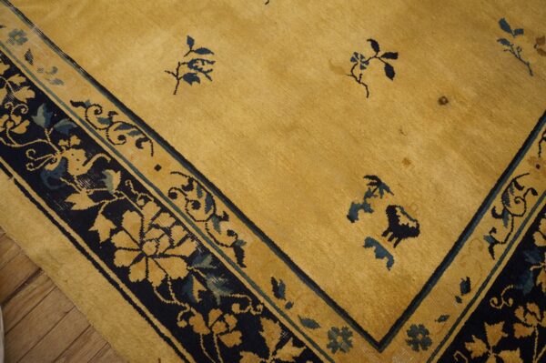 Gold rug corner showing a dark blue floral border and scattered stylized motifs in the field over wood floor.