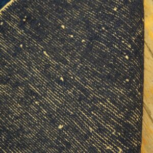 Detail of a rug underside showing dark blue weaving with light beige diagonal striations on wood flooring.