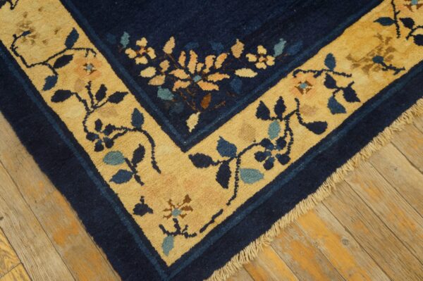 Close-up of a navy blue rug corner showing a golden yellow border with intricate blue and pink floral patterns on wood flooring.