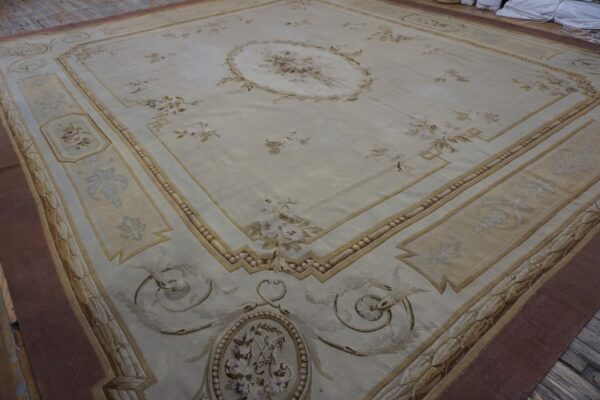 Large pale ivory rug with soft floral details and tan classical borders rests on weathered wood.