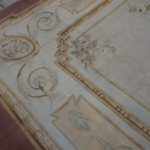 Ornate low-pile rug featuring pale blue, gold, and mauve floral designs over worn wood flooring.