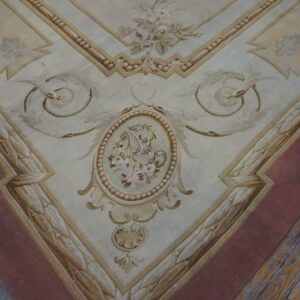 Cream and gold low-pile rug with elaborate neoclassical designs and a wide mauve border on rustic wood flooring.