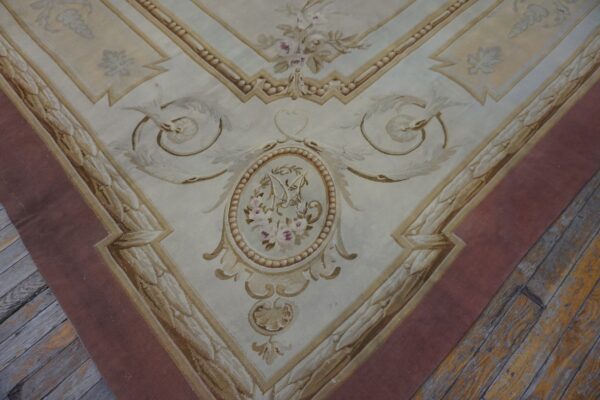 Cream and gold low-pile rug with elaborate neoclassical designs and a wide mauve border on rustic wood flooring.