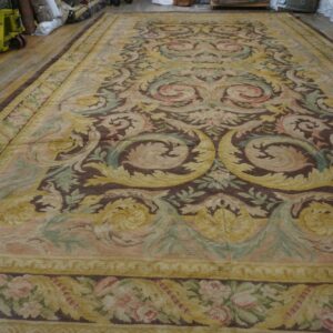 Low-pile tapestry rug with brown, yellow, pink, and sage scrollwork covering a wide-plank wood floor.