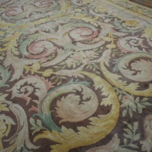 Ornate low-pile rug detailed with scrolling acanthus leaves in pale yellow, pink, and green on a brown background.