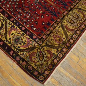 Detailed corner of a red rug with a gold floral border, set against worn, yellowish wood flooring.