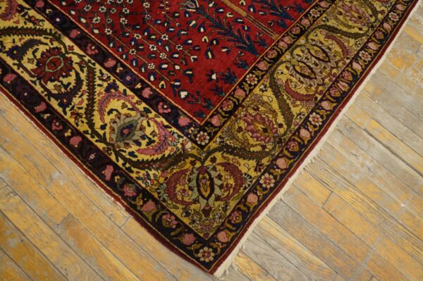 Detailed corner of a red rug with a gold floral border, set against worn, yellowish wood flooring.