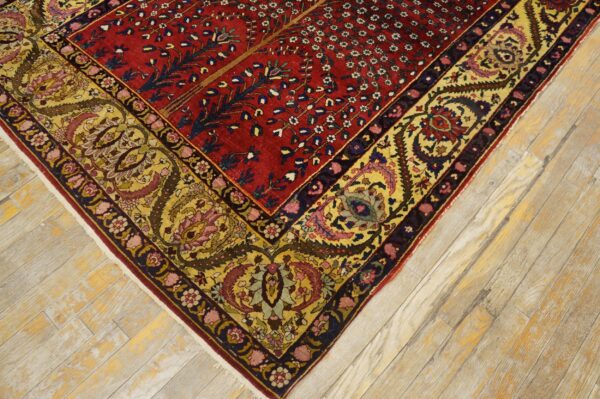 Ornate red field rug with a detailed gold and yellow floral border rests on light wood planks.